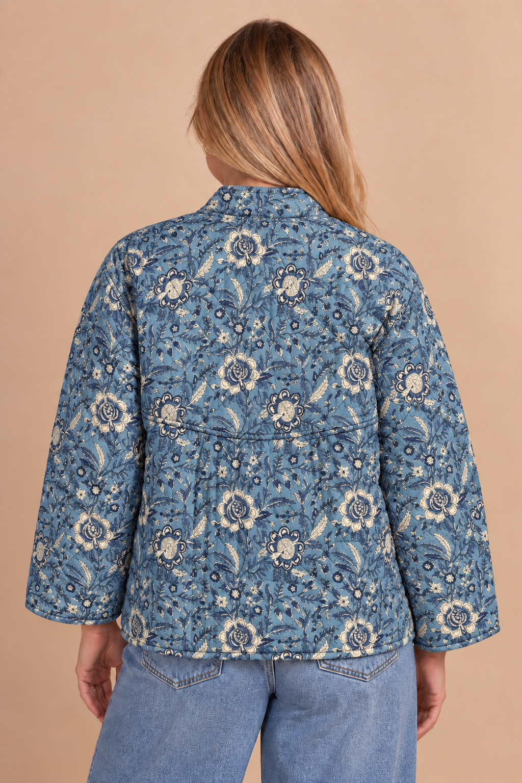 A woman viewed from the back wearing a blue quilted jacket with a cream and navy floral paisley pattern, paired with light blue jeans, against a solid beige background.