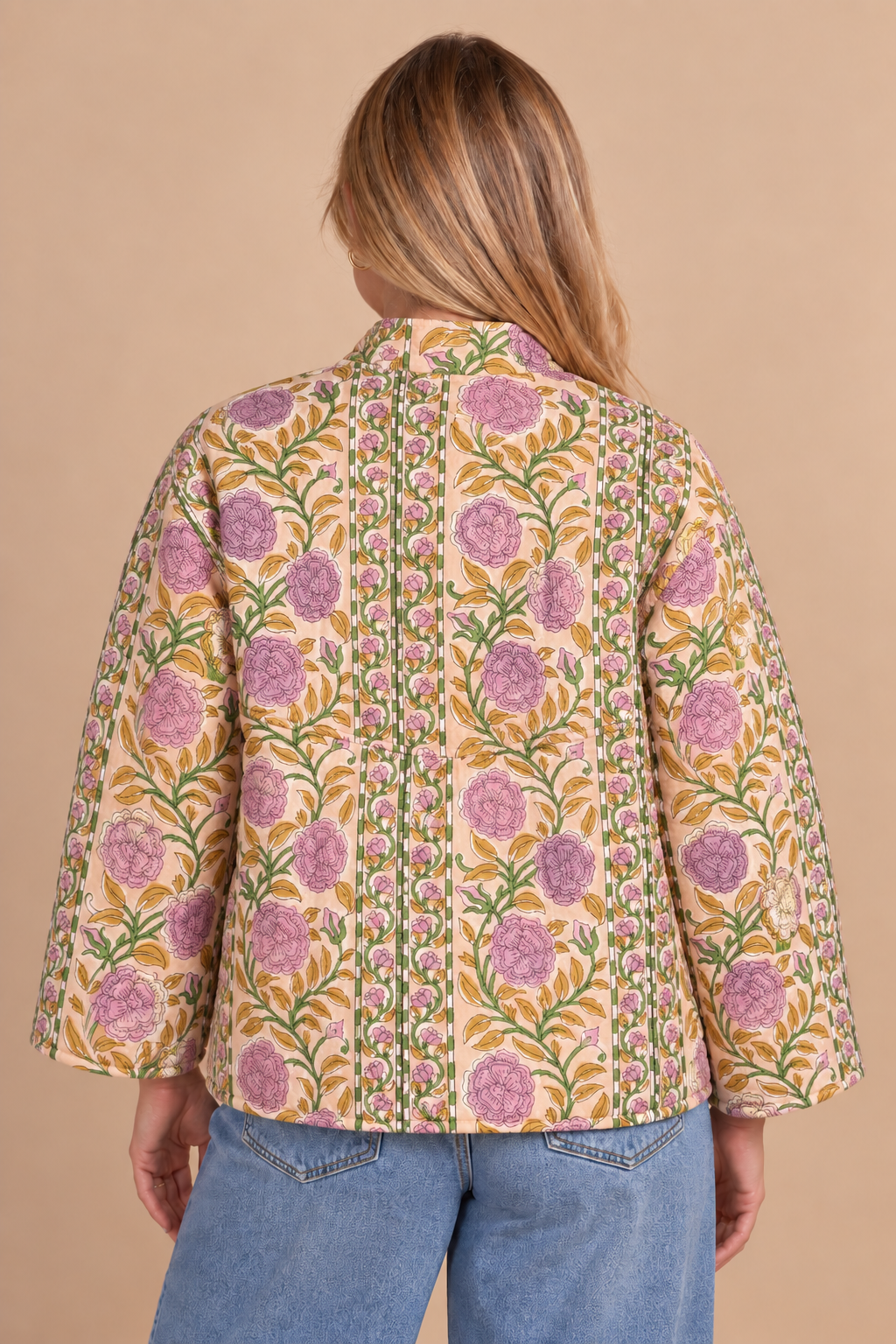 A woman shown from the back wearing a mustard yellow floral jacket with purple flowers and green leaves, paired with blue jeans, against a solid beige background.