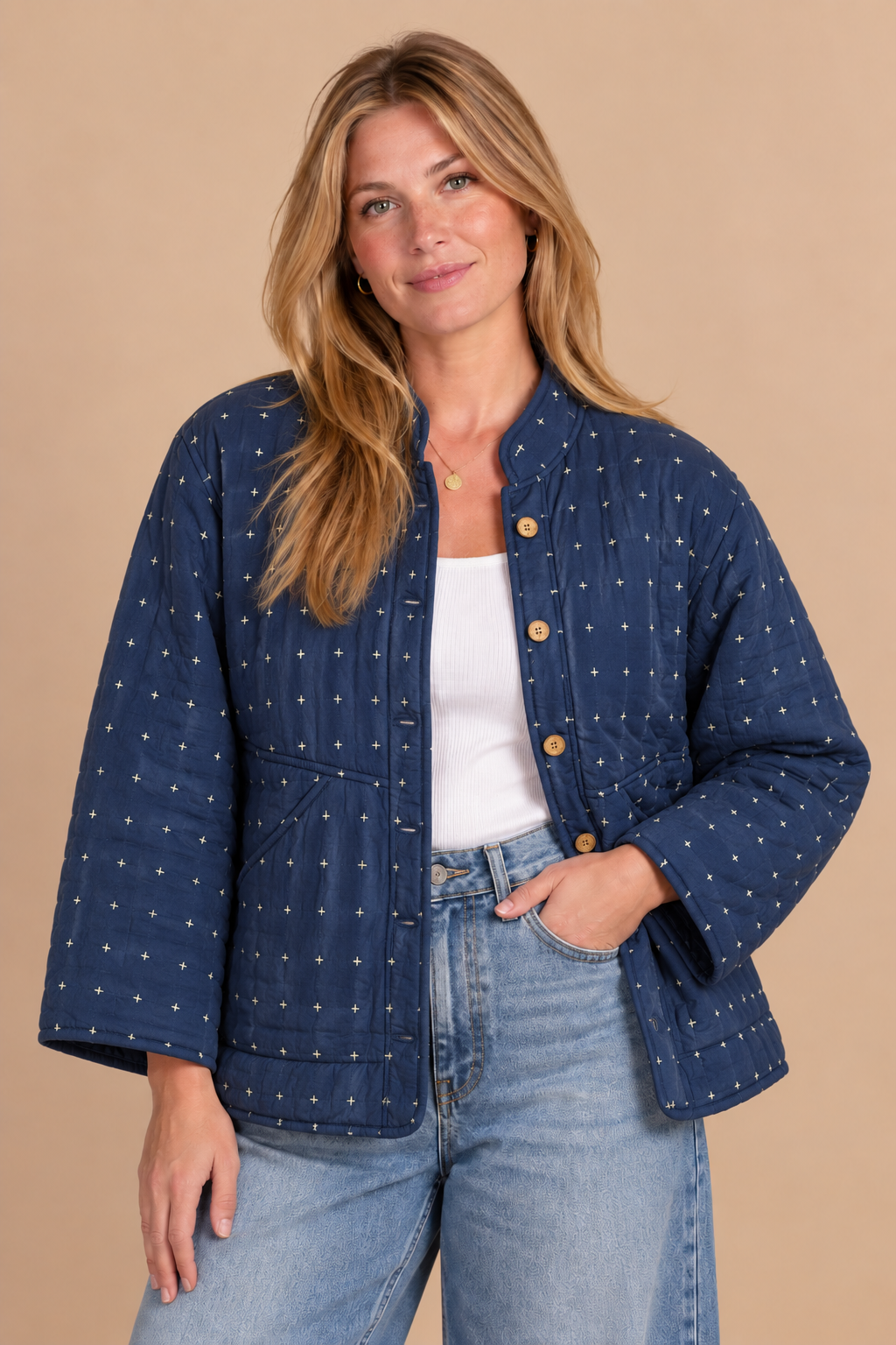 A woman modeling a navy blue quilted jacket with a small white polka dot pattern, styled over a white tank top and light blue jeans against a solid beige background.