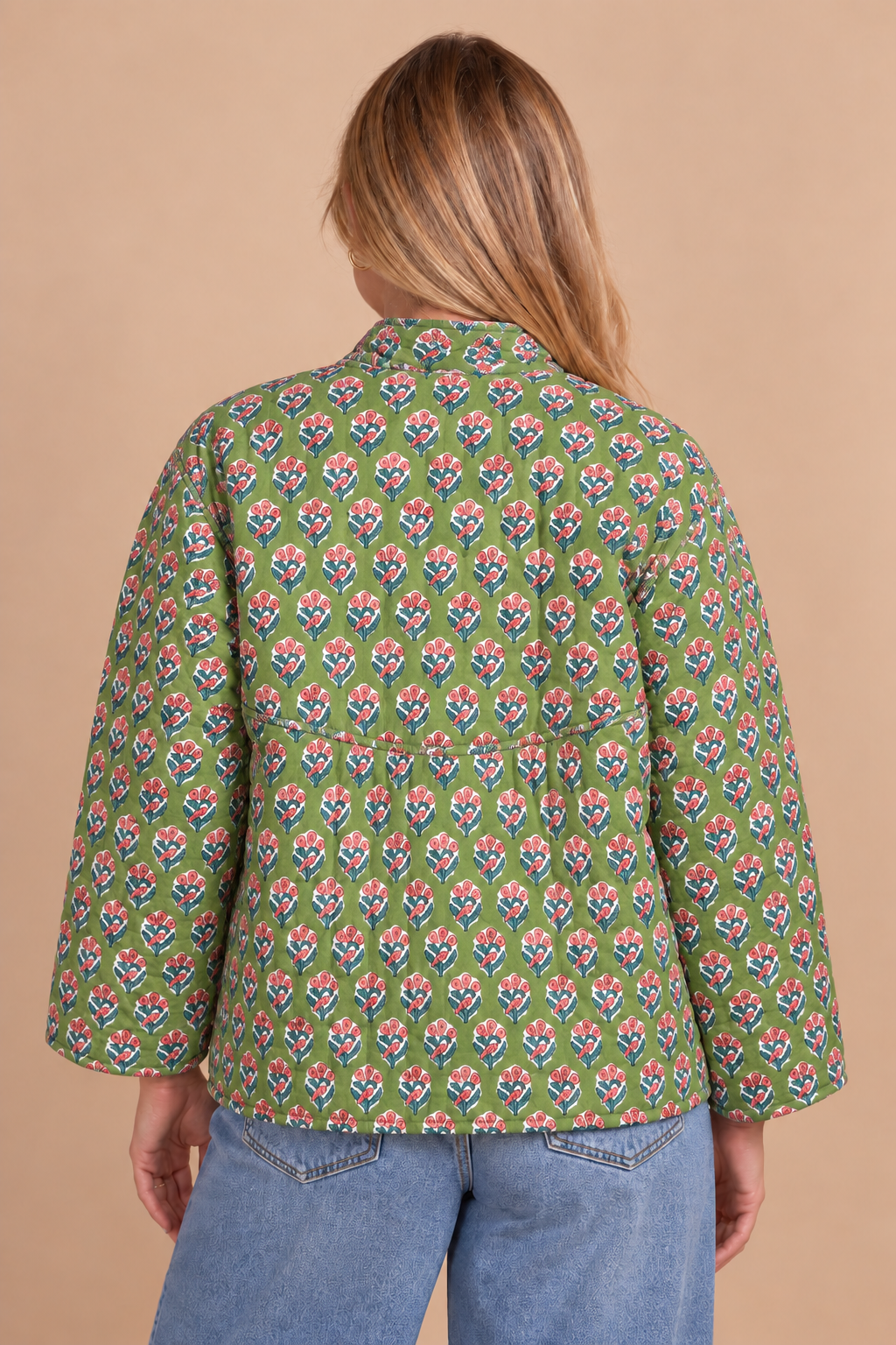 A woman viewed from the back wearing a green long-sleeve button-up shirt with a colorful floral block print, paired with blue jeans, against a solid beige background.