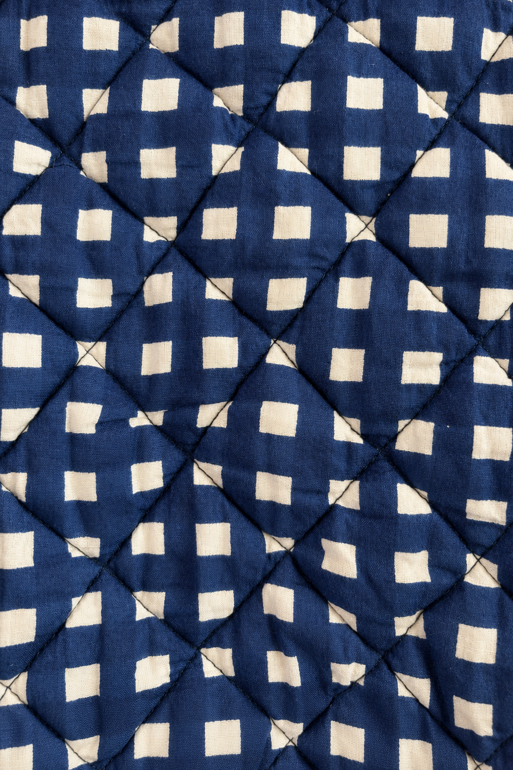 A close-up of a quilted fabric with a repeating cream-colored square pattern on a deep navy blue background, stitched diagonally in a diamond grid.
