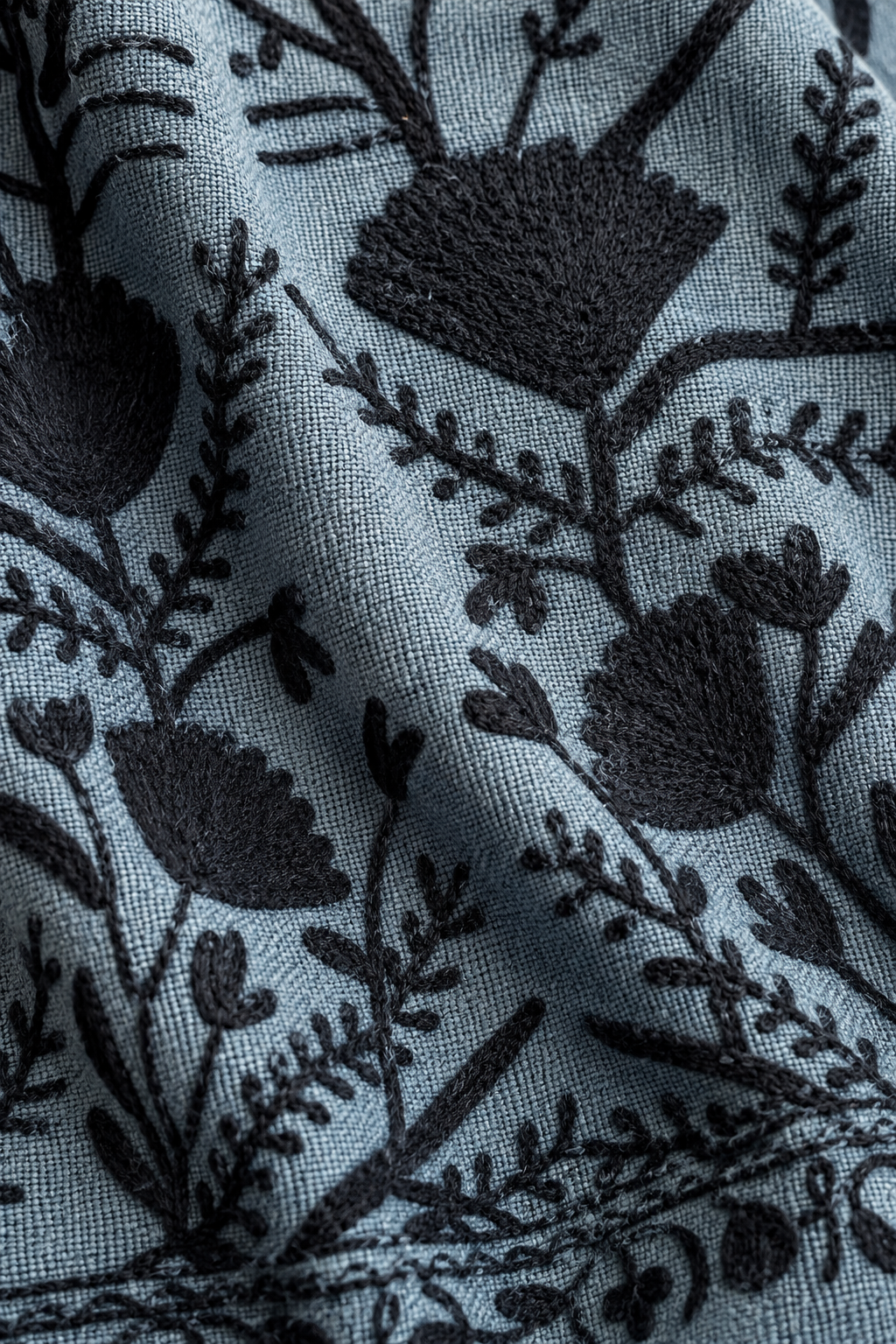 Close-up of a fabric surface with a dark floral and botanical embroidered pattern on a muted blue-gray woven background.