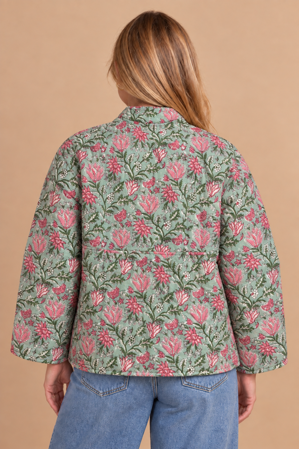 A woman viewed from the back wearing a light sage green jacket with a pink and white floral pattern, paired with blue jeans, against a solid tan background.