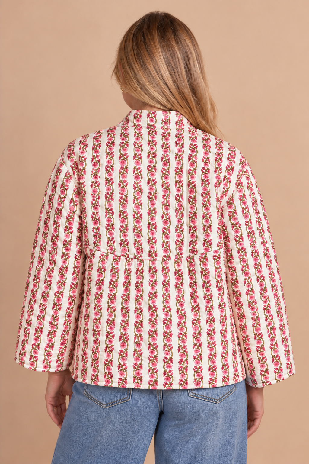 A woman viewed from the back wearing a white three-quarter sleeve blazer with vertical red and pink floral stripes, paired with blue jeans, against a solid beige background.