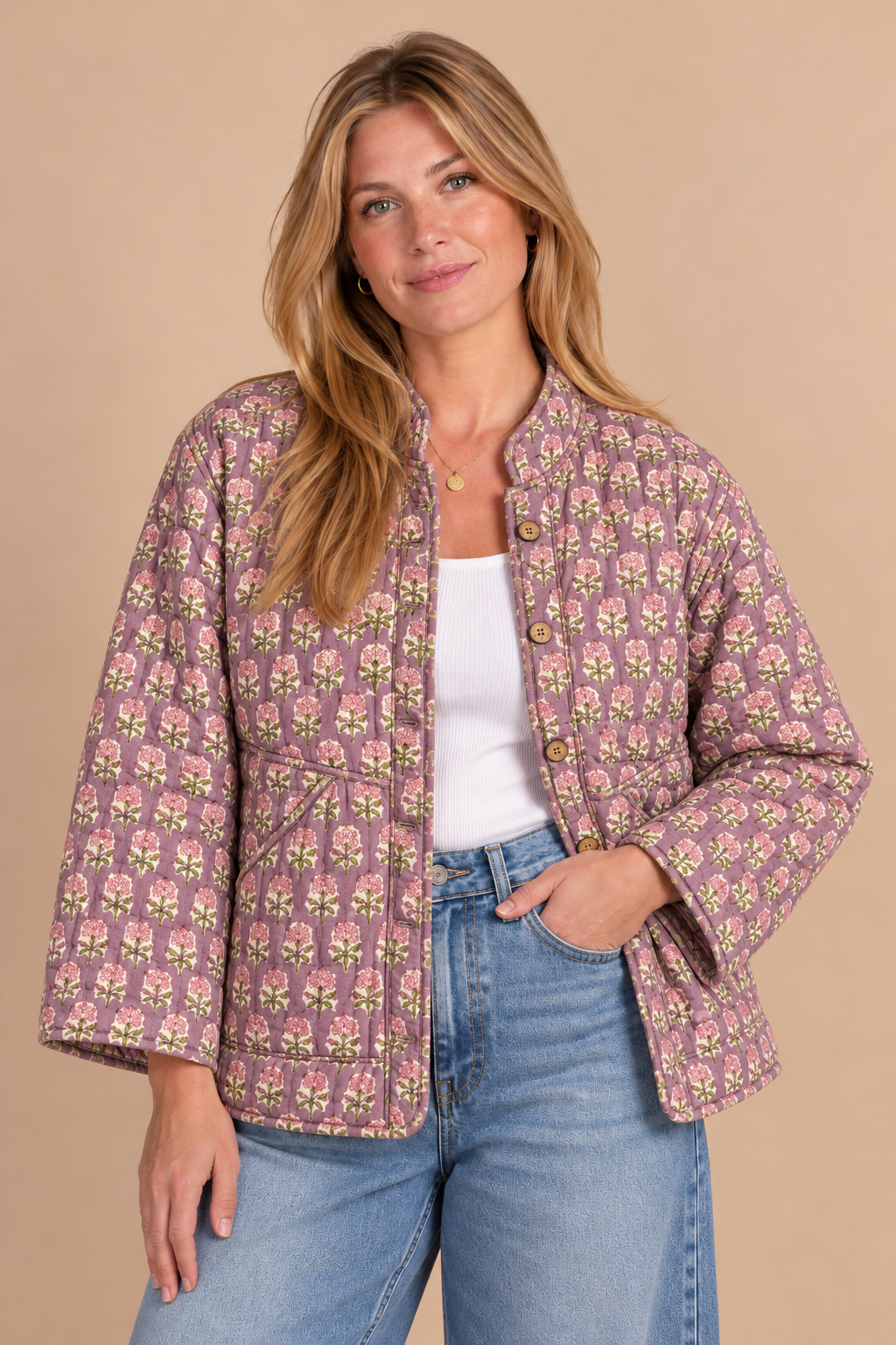 A woman modeling a cropped, quilted jacket with a pink and green floral pattern, worn over a white tank top and blue jeans against a solid beige background.