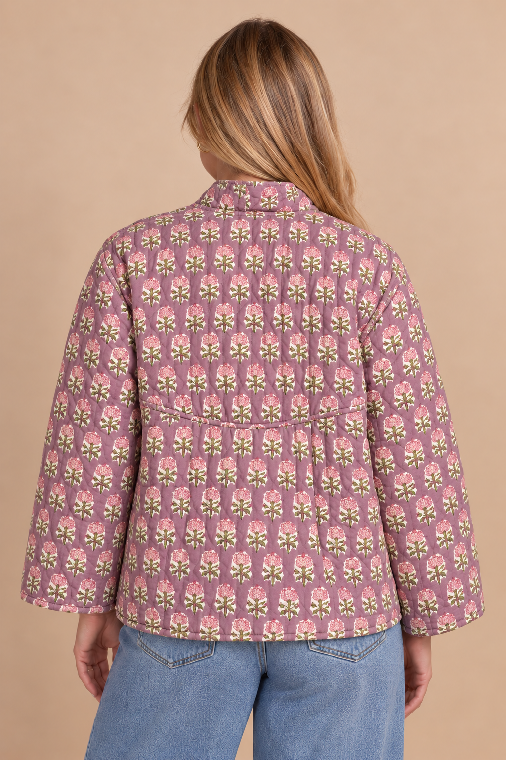 A woman viewed from the back wearing a pink quilted jacket with a repeating floral block print in green, yellow, and white, paired with blue jeans against a solid beige background.