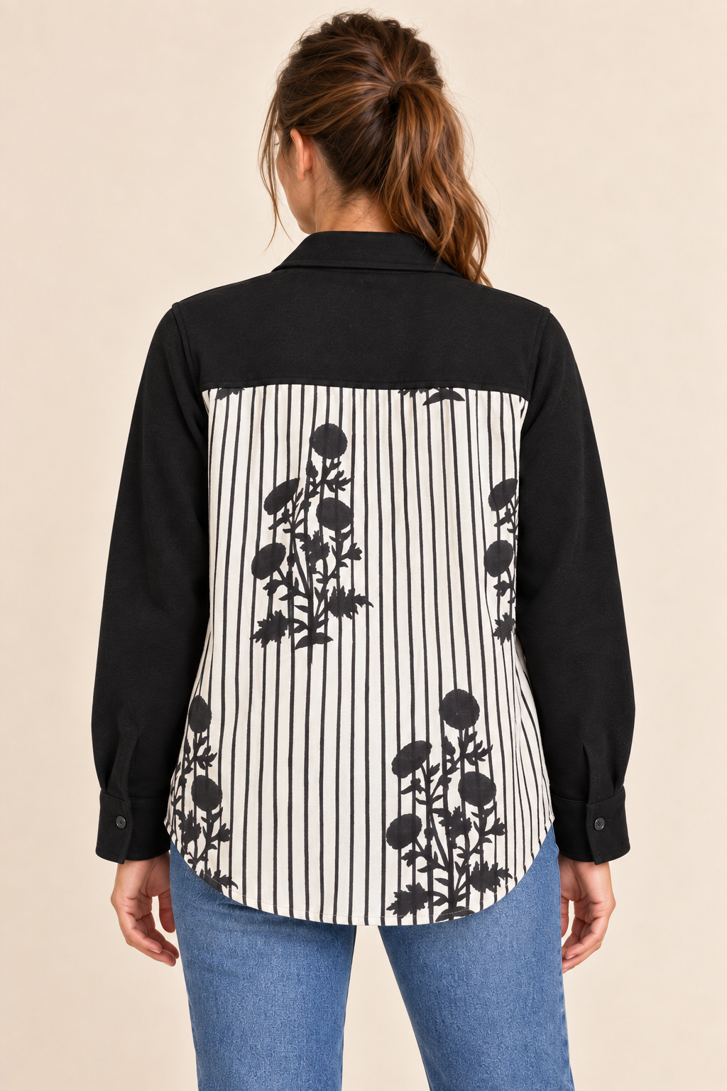 A woman viewed from the back wearing a long-sleeve button-up shirt with a black yoke and collar, paired with a black-and-white vertically striped body featuring black floral motifs, against a plain beige studio background.