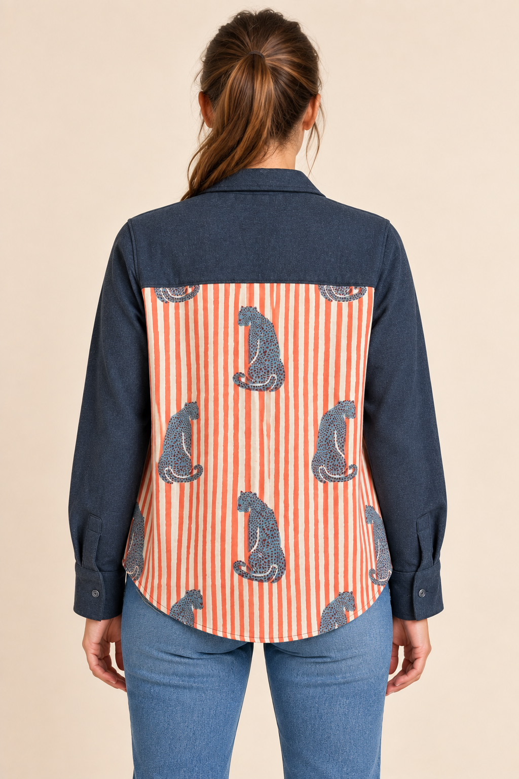 A woman viewed from the back wearing a long-sleeve button-up shirt with dark navy blue sleeves and shoulders, and a back panel featuring vertical orange and white stripes with gray cat silhouettes. She is also wearing blue jeans against a plain beige studio background.