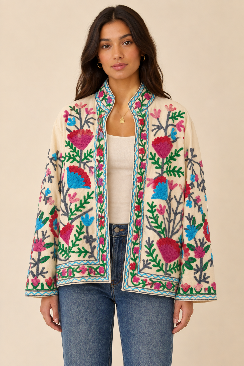 A woman modeling a cream-colored jacket with vibrant, multicolored floral and heart-shaped embroidery in pink, blue, green, and red, styled over a white tank top and blue jeans against a solid beige background.