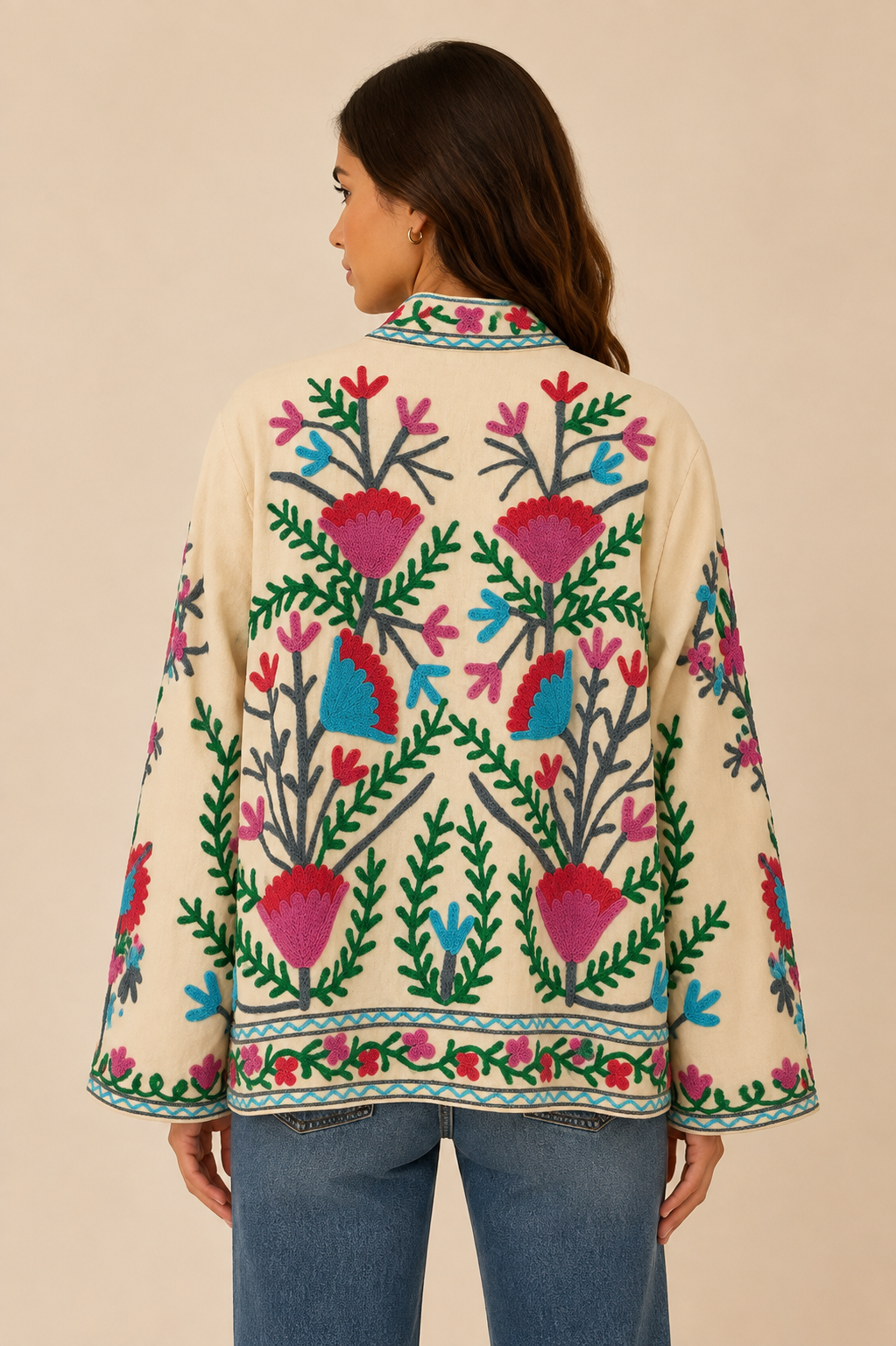 A woman shown from the back wearing a cream-colored jacket with vibrant, colorful floral and botanical embroidery, paired with blue jeans, against a solid beige background.