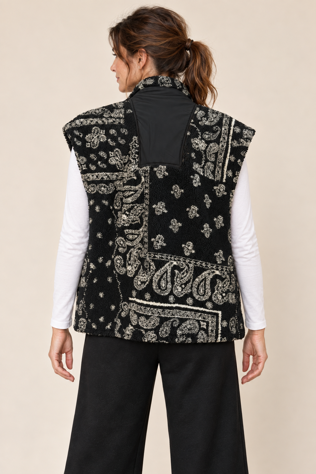 A woman shown from the back wearing a sleeveless black vest with a white paisley bandana print over a black top, paired with black pants, against a plain beige studio background.
