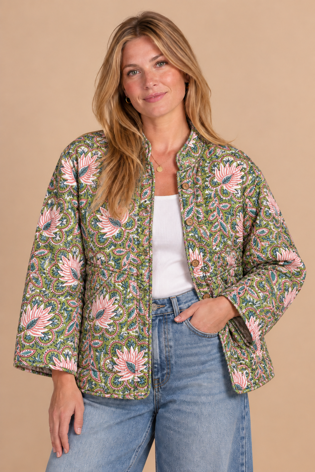 A woman modeling a cropped, long-sleeve quilted jacket with a green, pink, and blue floral pattern, worn over a white top and blue jeans against a solid beige background.
