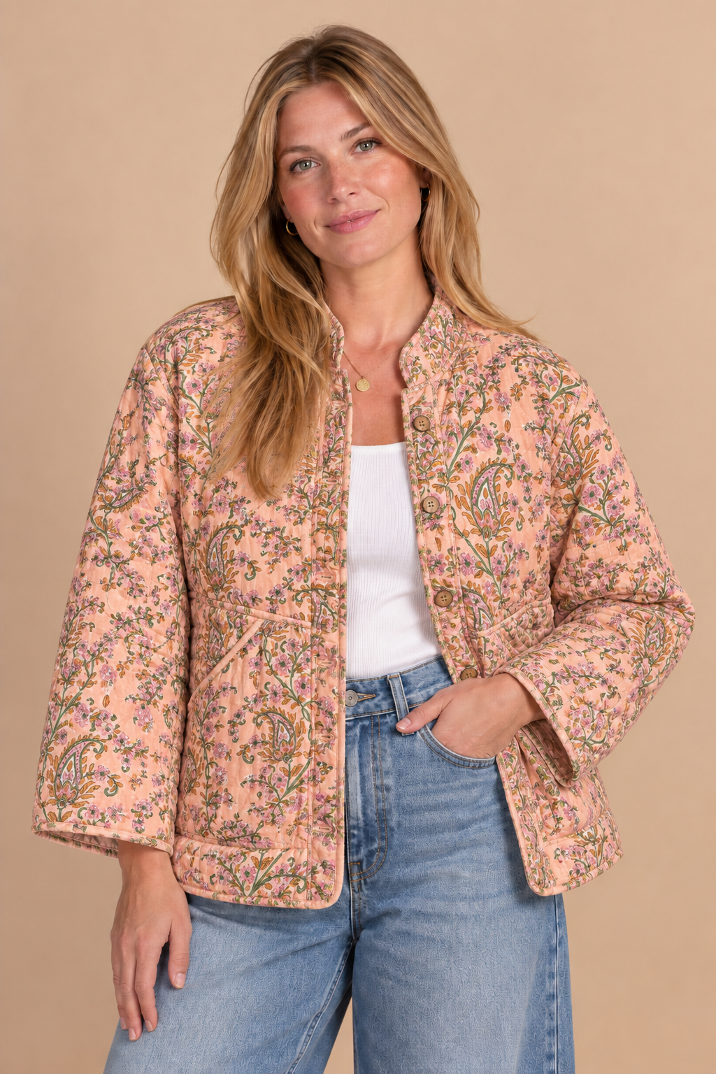 A woman modeling a peach-colored quilted jacket with a green and yellow paisley pattern, worn over a white top and blue jeans, against a solid beige background.