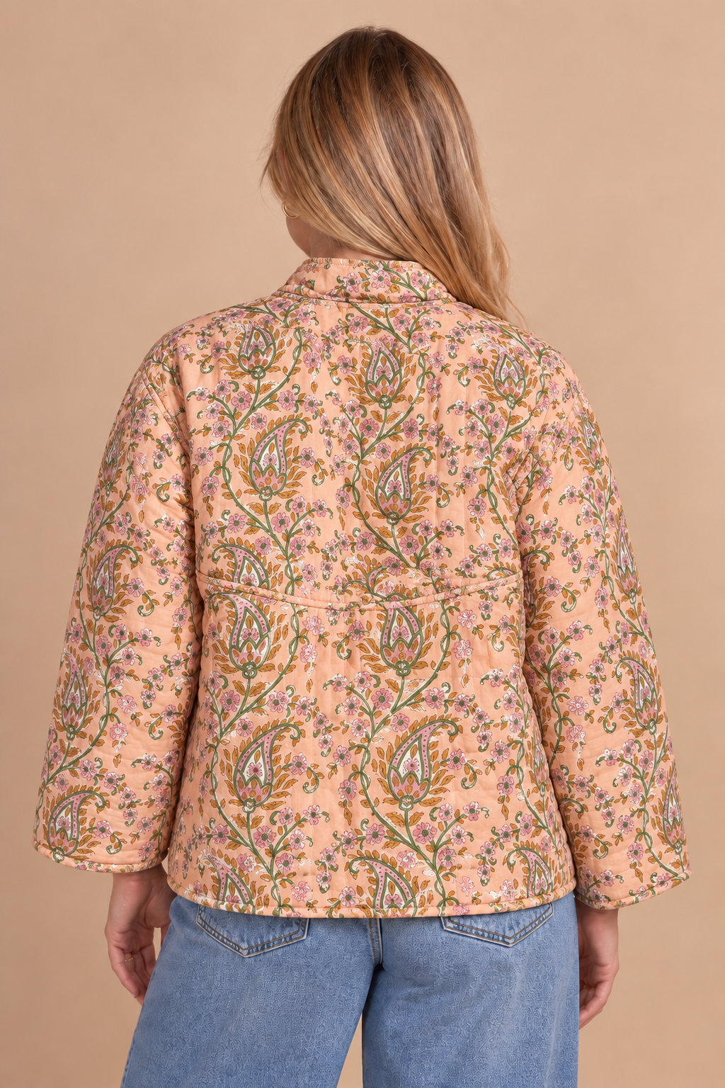 A woman viewed from behind wearing a peach-colored quilted jacket with a green and blue paisley pattern, paired with blue jeans, against a solid beige background.