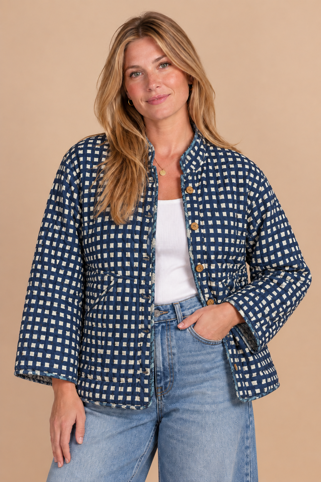 A woman modeling a navy and white gingham patterned jacket with a stand collar, button front, and flared sleeves, paired with a white top and light blue jeans against a solid beige background.