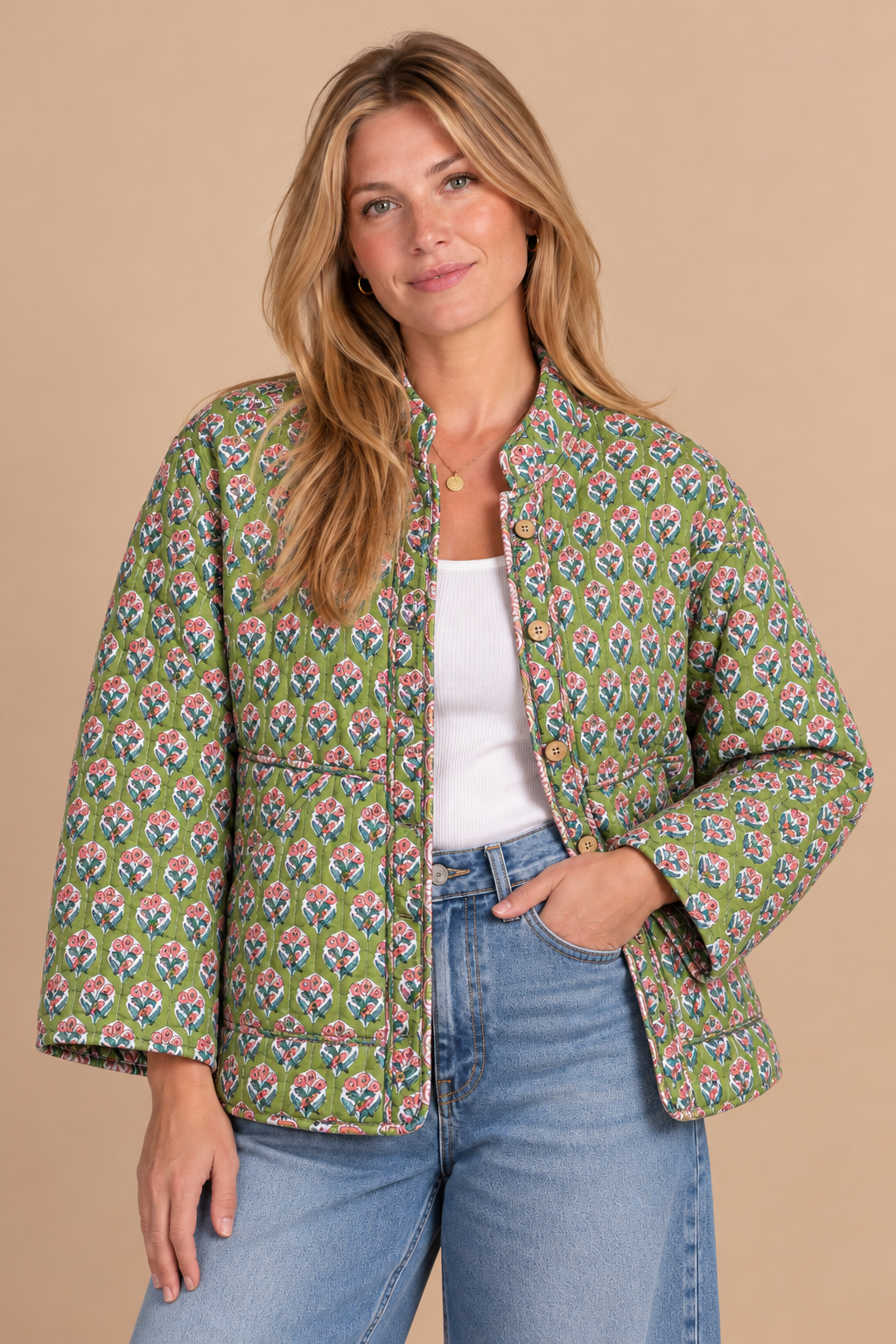 A woman modeling a green patterned quilted jacket with a white top and blue jeans, standing against a solid beige background.