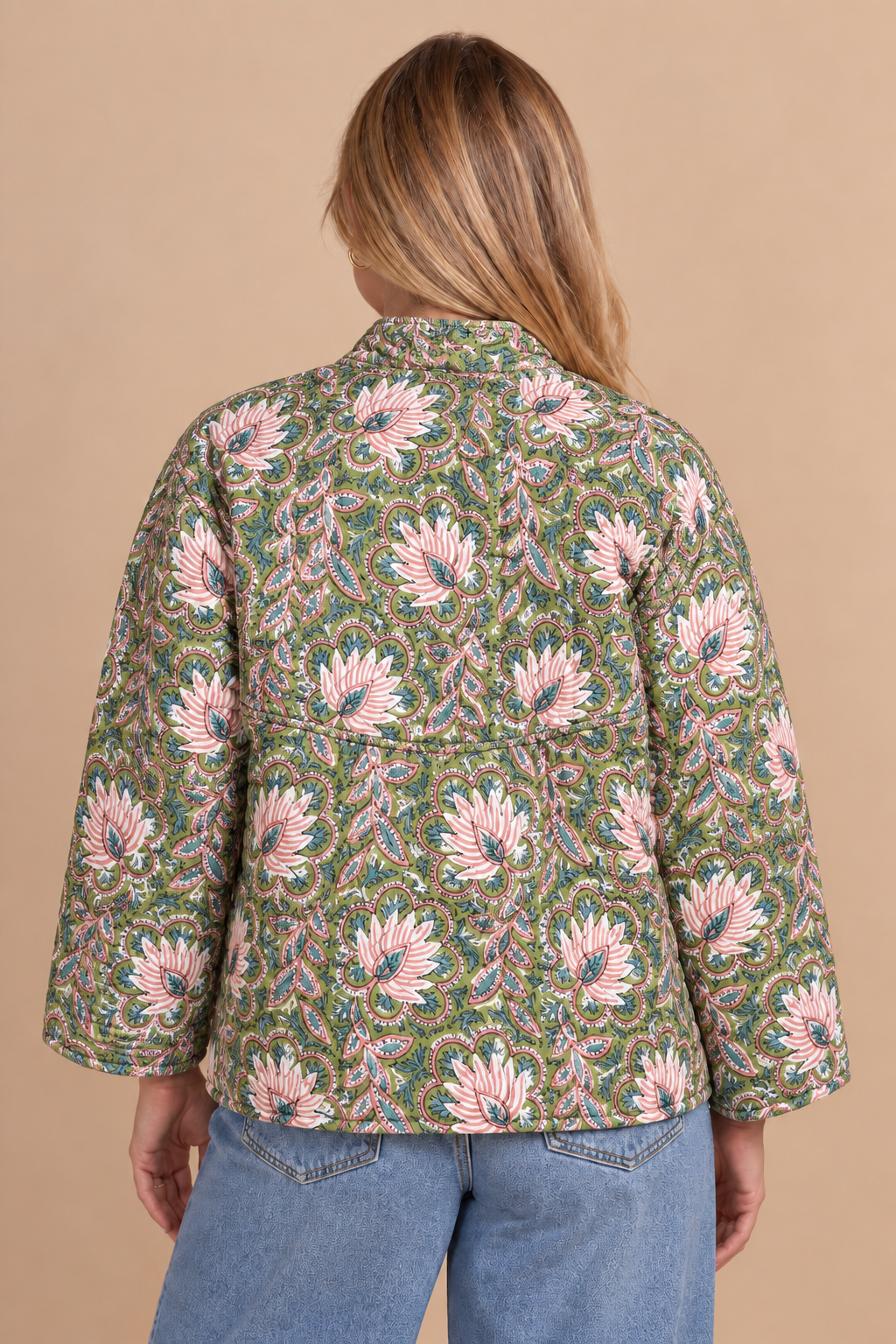 A woman viewed from the back wearing a green patterned jacket with pink and white floral motifs, paired with blue jeans, against a solid beige background.