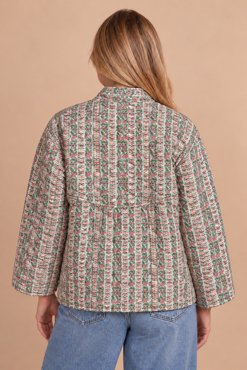 A woman viewed from the back wearing a long-sleeve button-front jacket with an intricate multicolor vertical pattern in green, pink, and beige, paired with blue jeans against a solid tan background.