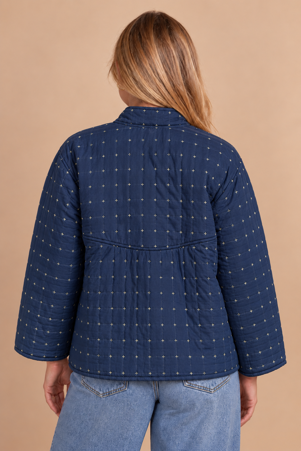 A woman viewed from the back wearing a navy blue quilted jacket with a small white geometric pattern, paired with light blue jeans, against a solid beige background.