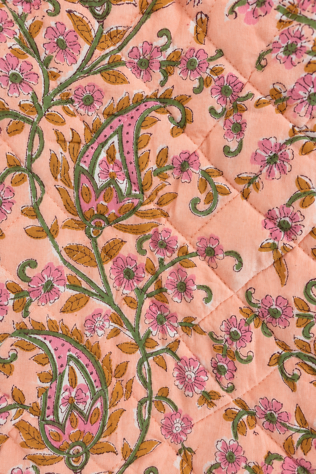 A close-up of a quilted fabric featuring a peach-pink background with a detailed block-print style floral and paisley pattern in green, brown, and pink.