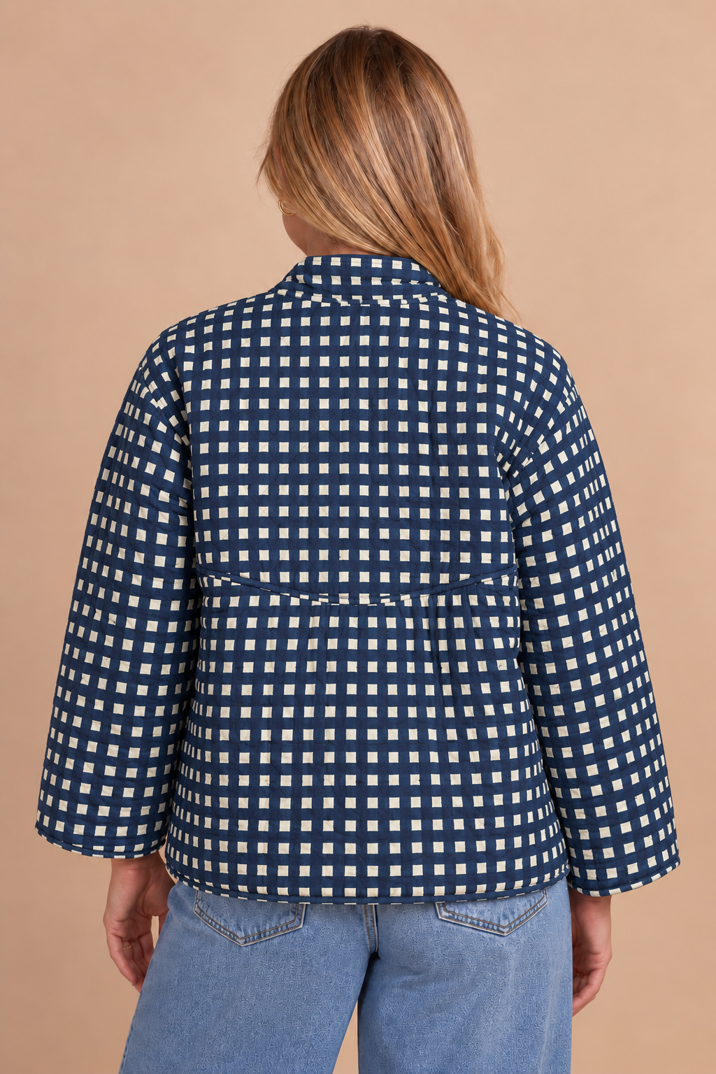 A woman viewed from the back wearing a navy blue and white gingham patterned jacket with three-quarter sleeves, paired with light blue jeans, against a solid beige background.