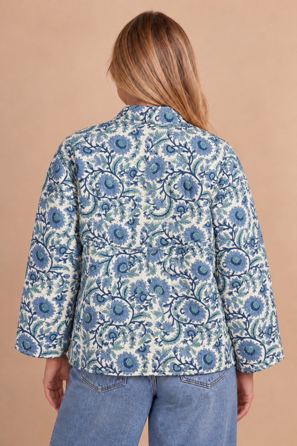 A woman viewed from the back wearing a blue and white floral patterned jacket over a light blue denim shirt, set against a solid beige background.