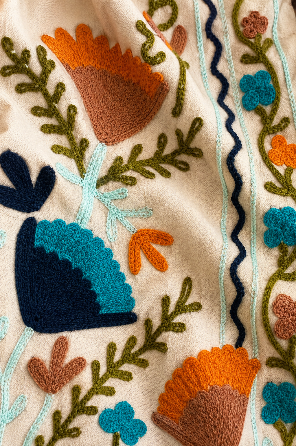 Close-up of a cream-colored textile featuring colorful hand-embroidered floral and leaf motifs in orange, teal, brown, green, and navy blue.