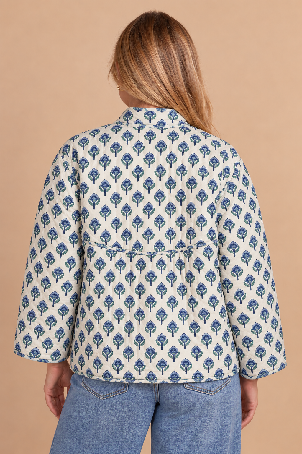 A woman viewed from the back wearing a cream-colored blouse with a blue geometric block print pattern and blue jeans, set against a solid beige background.