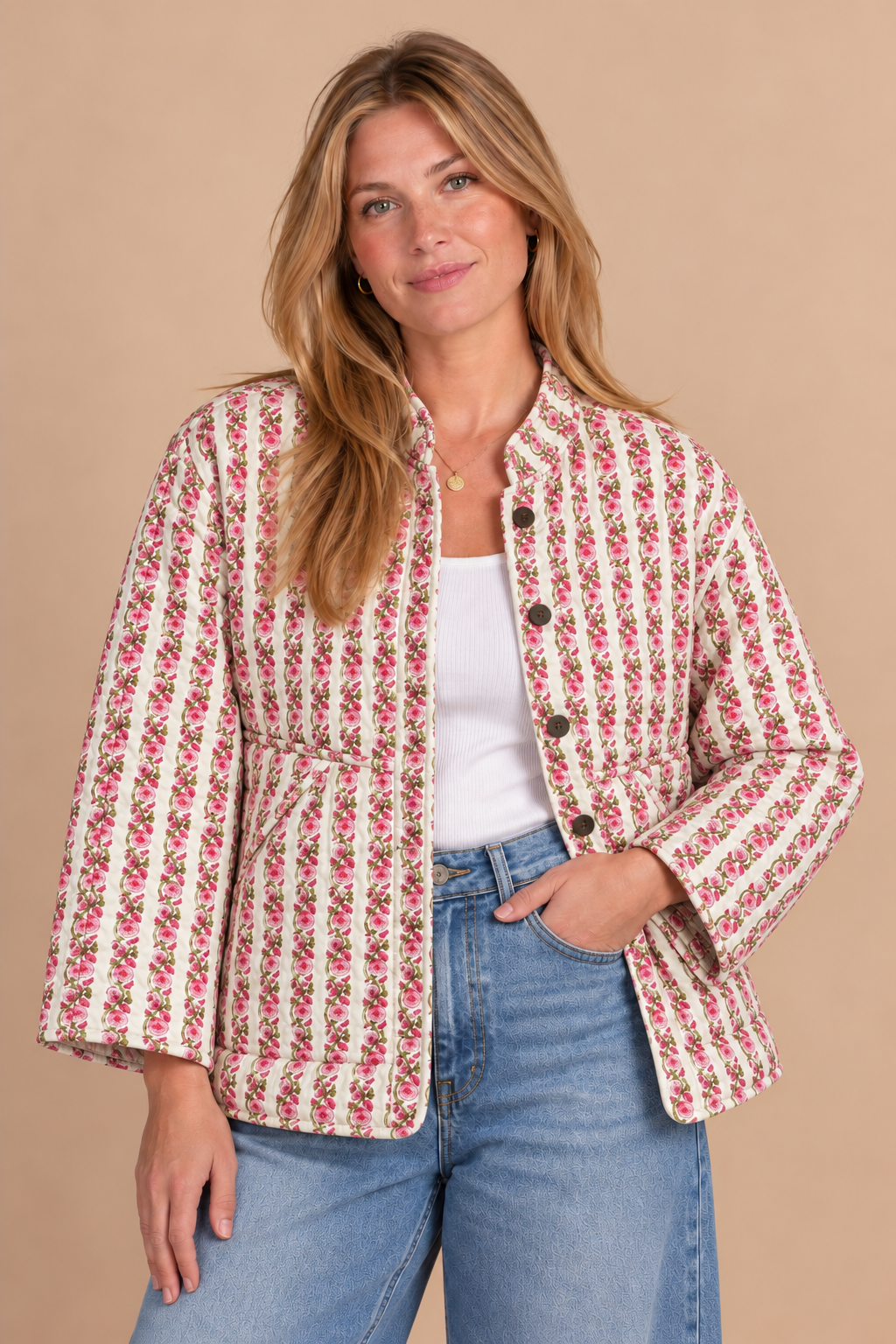 A woman modeling a cream-colored jacket with a pink and red floral pattern, worn over a white tank top and blue jeans, against a solid beige background.