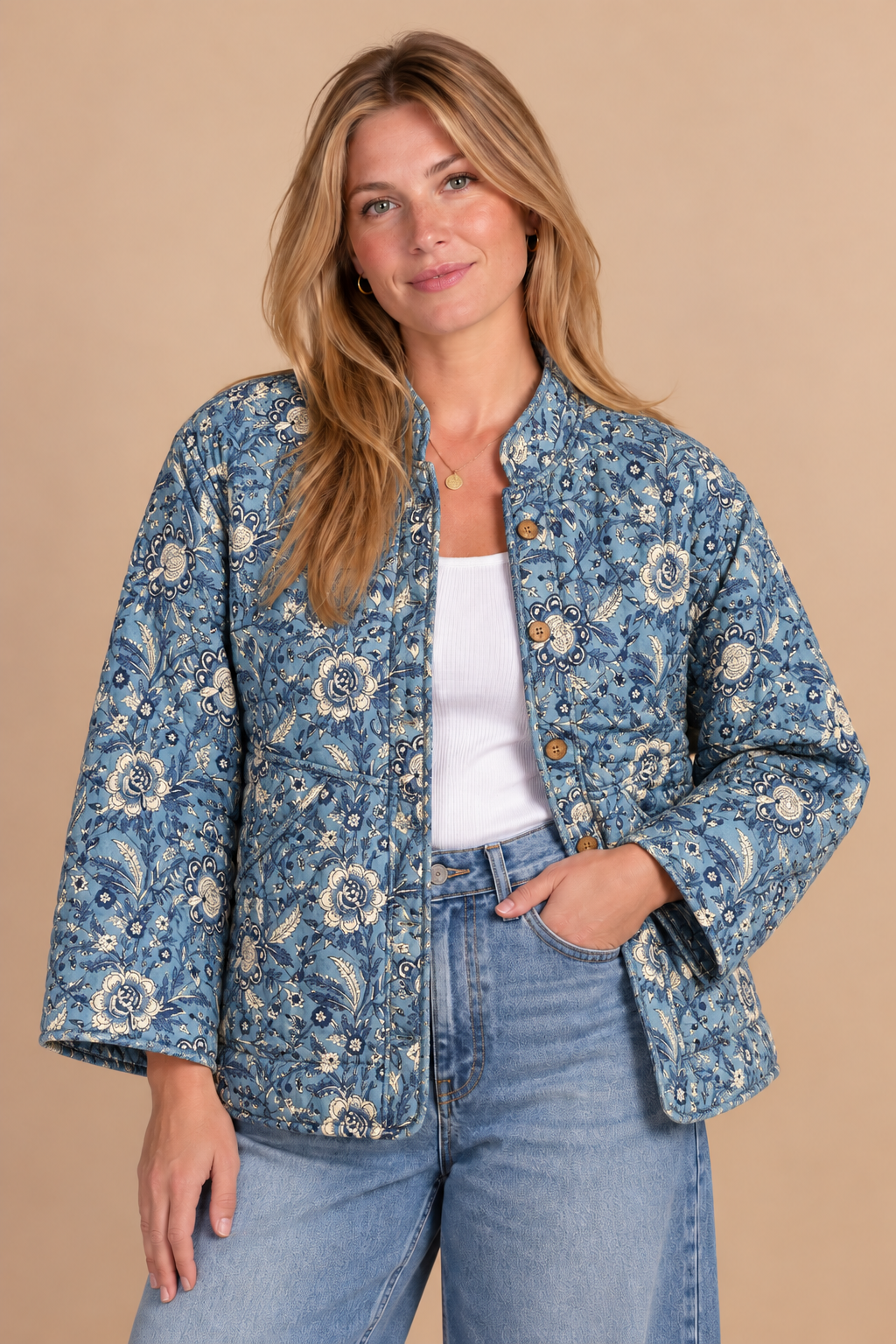A woman modeling a blue floral quilted jacket with a stand collar, button front, and three-quarter sleeves, paired with a white tank top and light-wash jeans against a solid beige background.