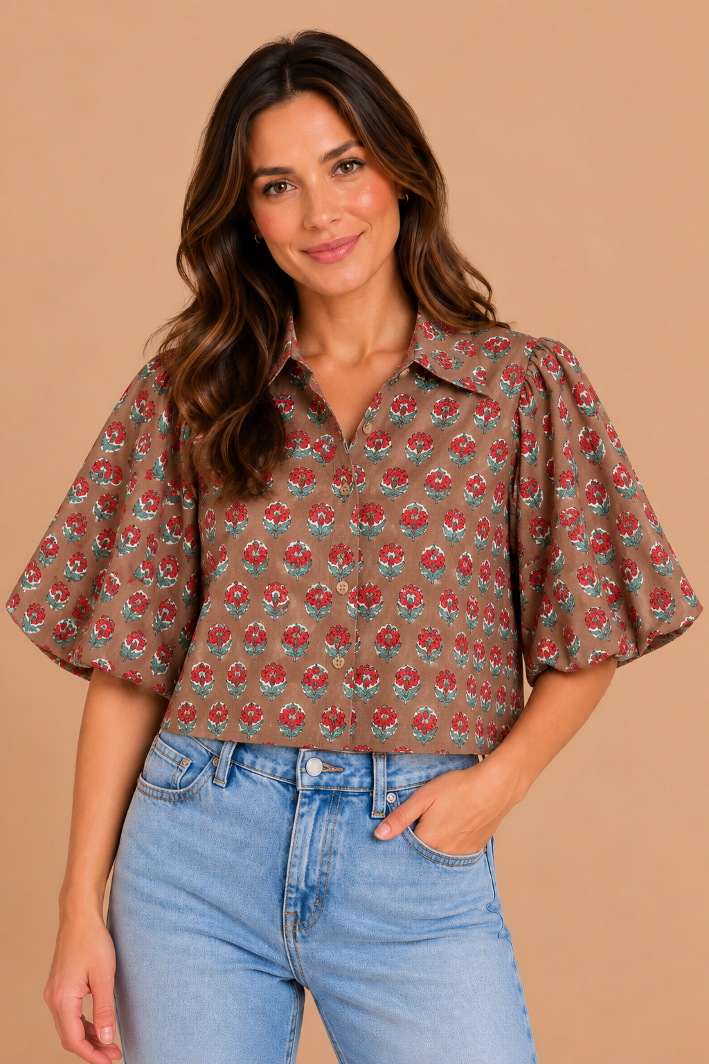 A woman wearing a brown short-sleeve button-up blouse with a red and green floral pattern, paired with light blue jeans, set against a solid beige background.