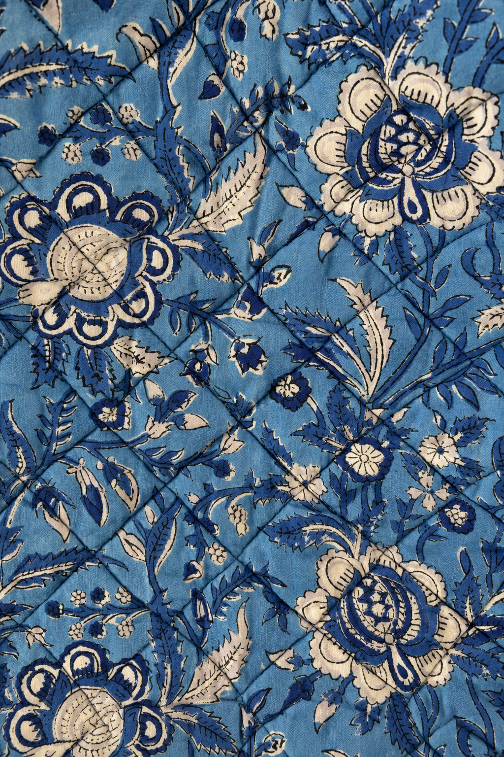 A close-up of a blue fabric with a vintage floral block print pattern in cream and dark blue, showing a quilted texture with diagonal stitching.