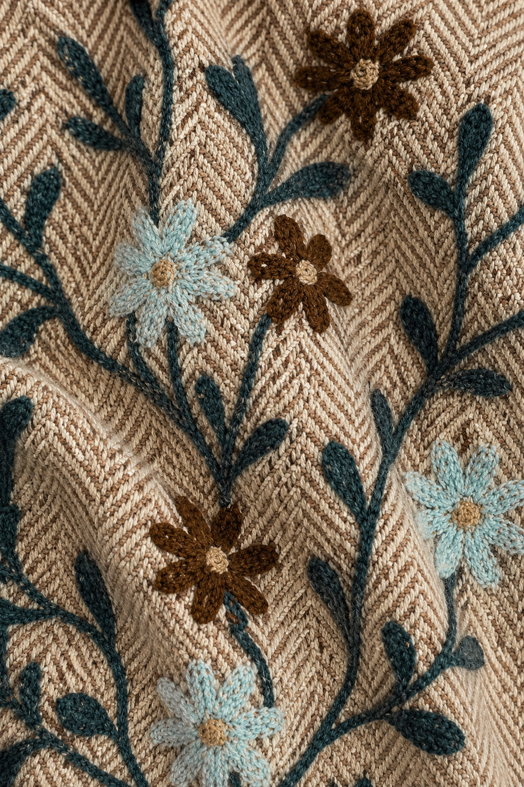 Close-up of a textured knit fabric with a beige herringbone pattern, decorated with embroidered floral vines in dark teal, brown, and light blue.