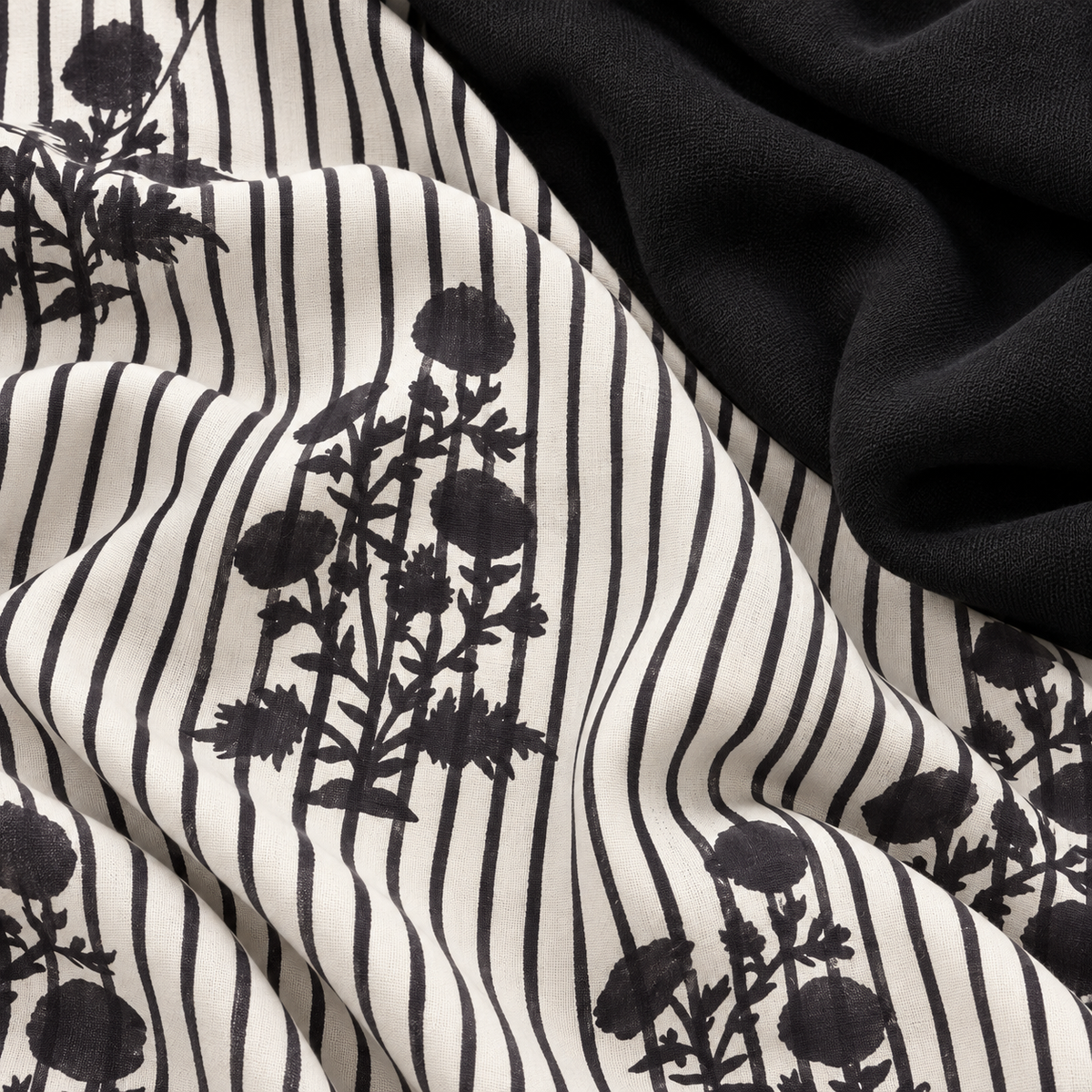 A close-up of draped fabric with a black and off-white vertical stripe pattern overlaid with black floral silhouettes, alongside a solid black fabric section.