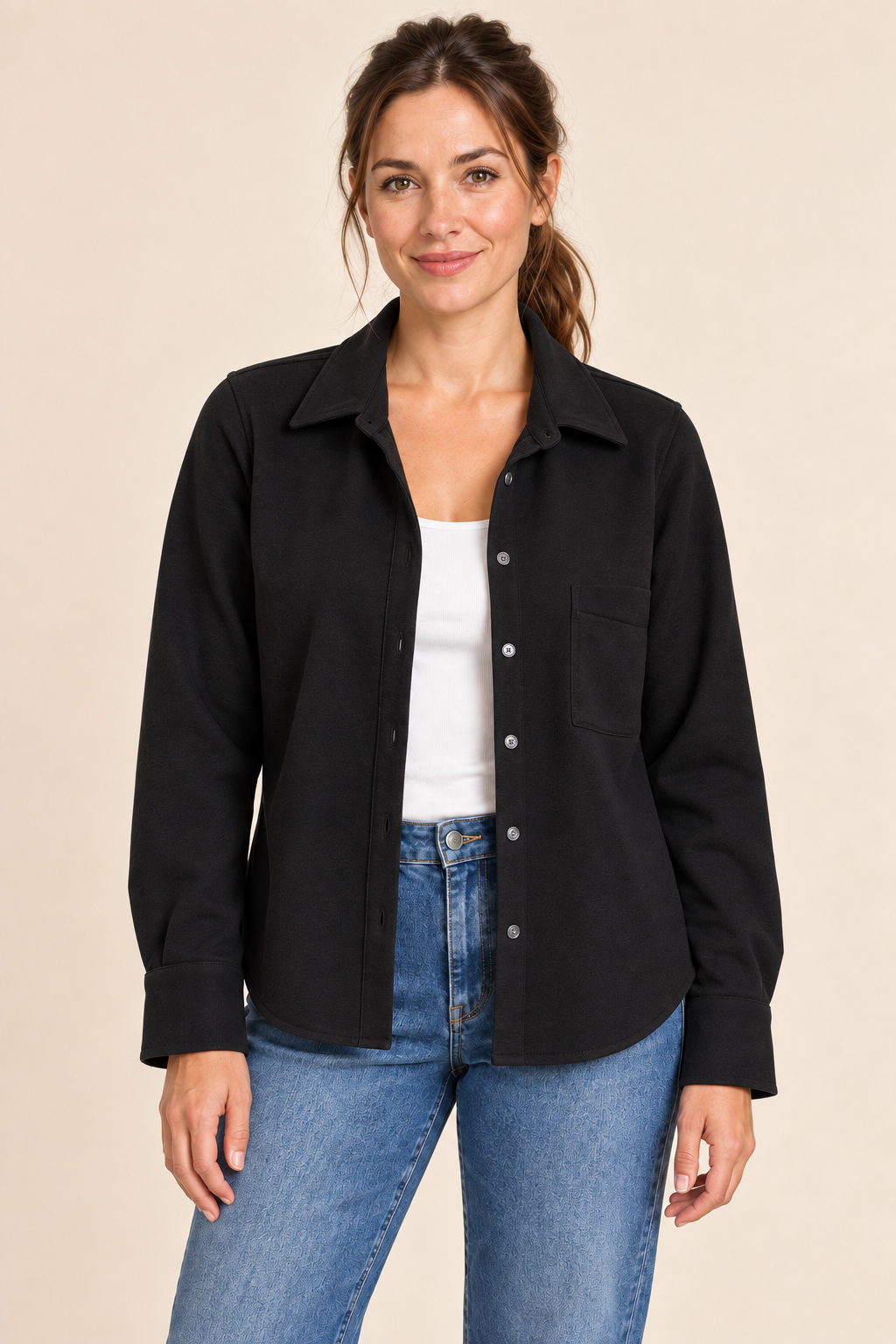 A woman modeling a black long-sleeve button-up shirt jacket over a white tank top and blue jeans, photographed against a plain beige studio background.