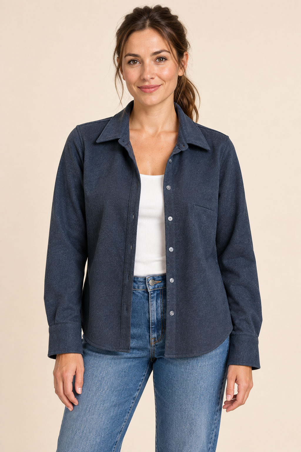 A woman modeling a dark blue long-sleeve button-up shirt over a white tank top and blue jeans, standing against a solid beige background.